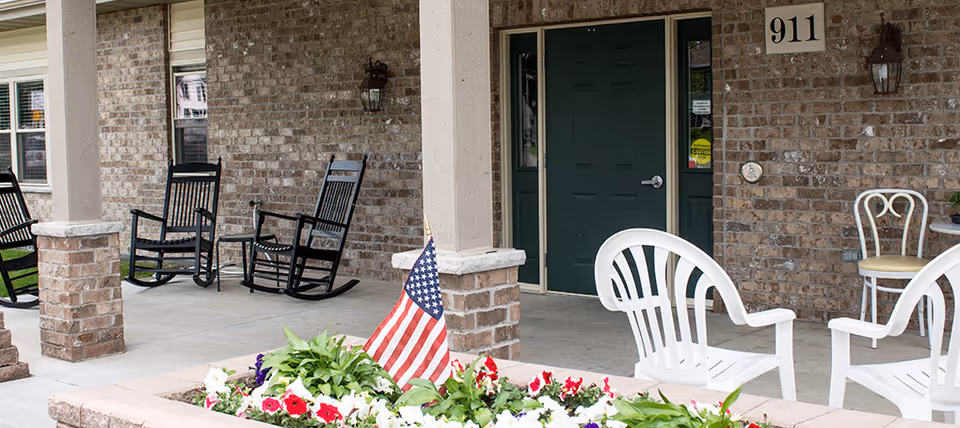 Front porch of a brick senior living facility with rocking chairs and flowers.