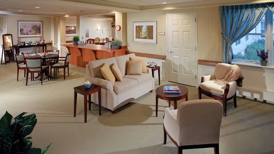 A cozy living room area in a senior living facility featuring a beige loveseat with multiple cushions, two armchairs, a round coffee table with books, and side tables. The room has a large window with blue curtains, a door, framed artwork on the walls, and a dining area with a table and chairs in the background. The space is warmly lit and decorated with plants and a mirror on the far wall.