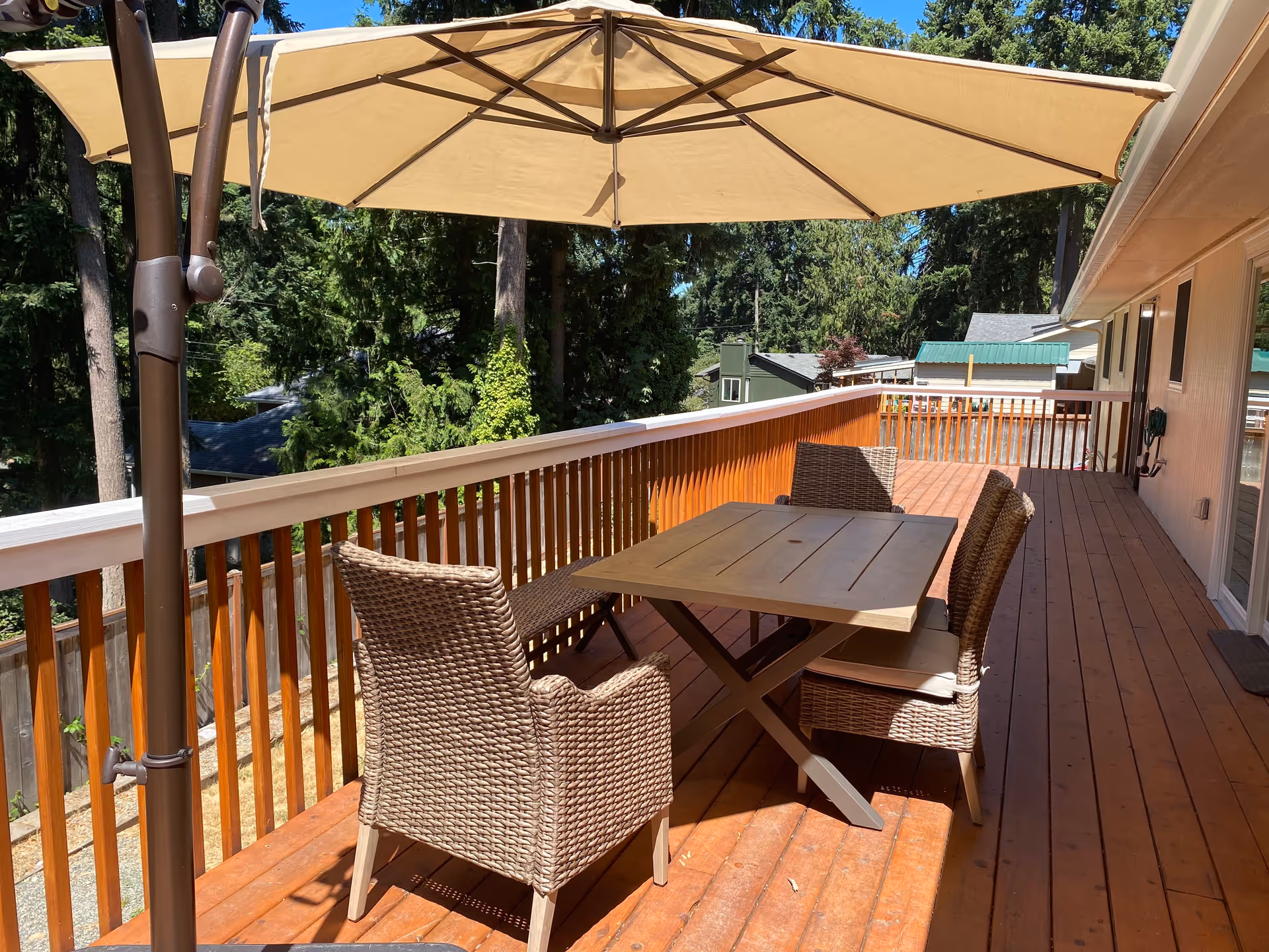 Outdoor wooden deck with a table and four wicker chairs under a large beige patio umbrella, surrounded by trees and neighboring houses.