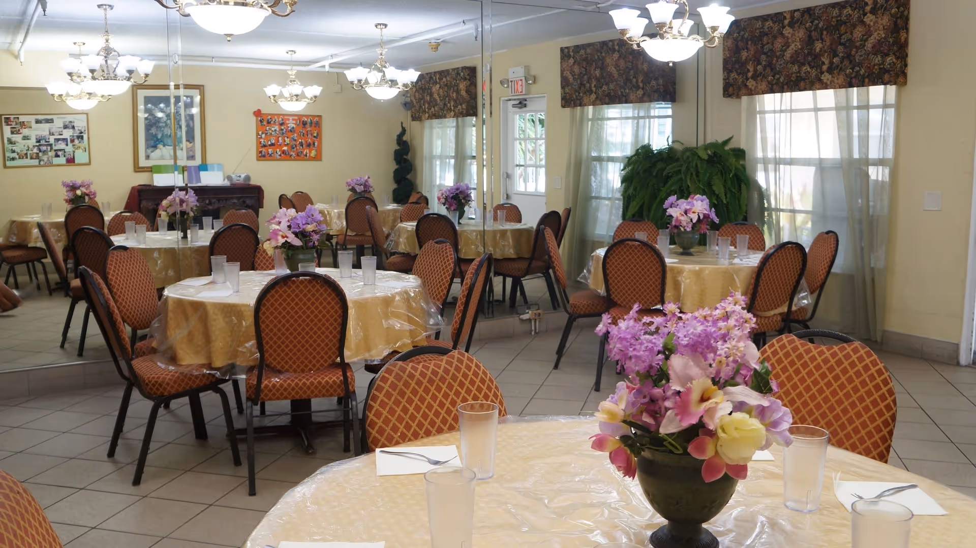 A dining room with multiple round tables covered with yellow tablecloths and clear plastic covers. Each table has a floral centerpiece with purple and pink flowers. The chairs have a red and gold diamond pattern. The room has large mirrors on one wall, floral valances over the windows, and several chandeliers hanging from the ceiling. There are also framed pictures and a bulletin board on the far wall.