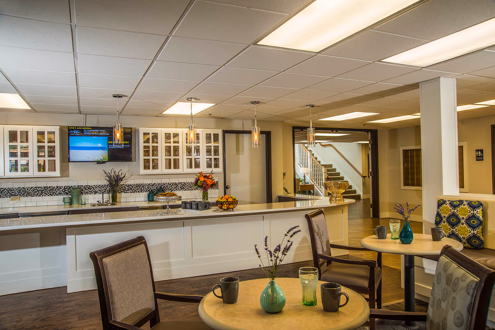 Communal dining and kitchenette area with a long white counter, pendant lights, small round tables with chairs, and a staircase in the background.