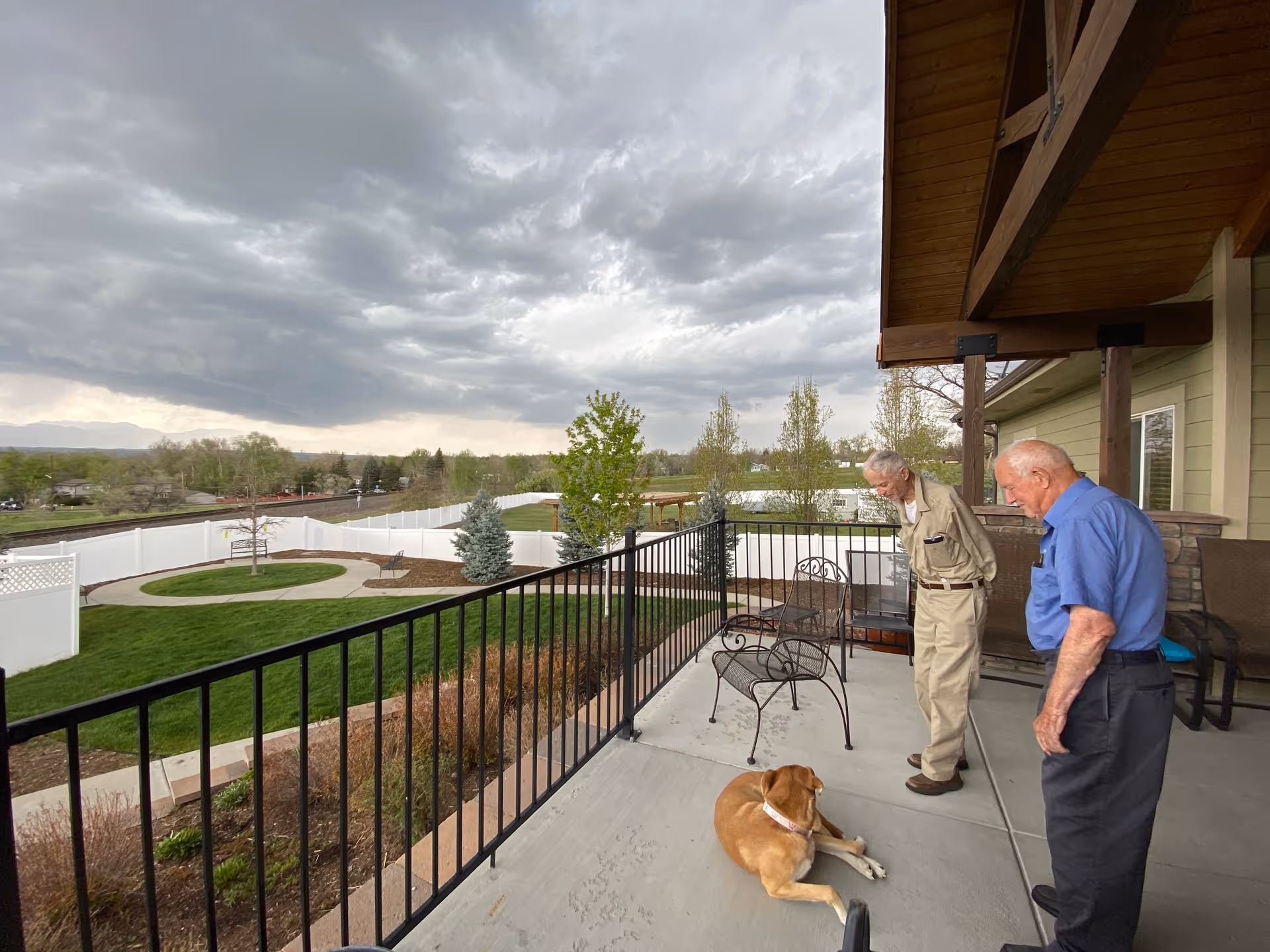 Two elderly men stand on a covered patio looking down at a dog lying on the concrete, with a fenced yard and cloudy sky beyond.