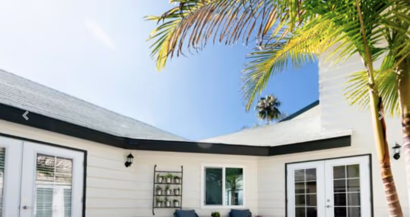 Outdoor patio area of a senior living facility with white walls, black trim, two sets of glass double doors, a small wall-mounted shelf with potted plants, and palm trees providing shade under a clear blue sky.