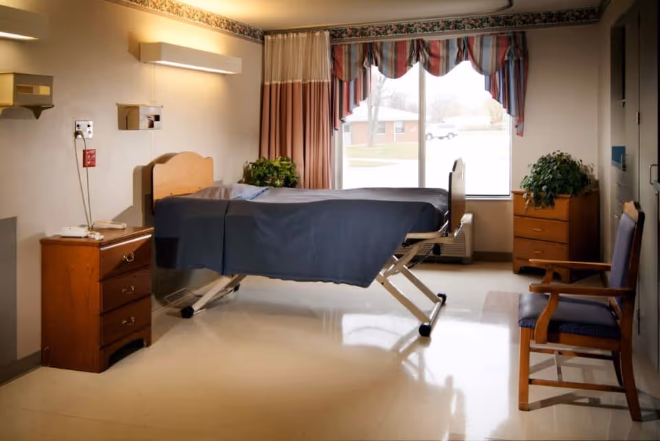 A healthcare facility room with a hospital bed covered in a dark blanket, a wooden nightstand with a telephone, a wooden dresser with a potted plant, a wooden chair with blue upholstery, and a large window with striped curtains letting in natural light.