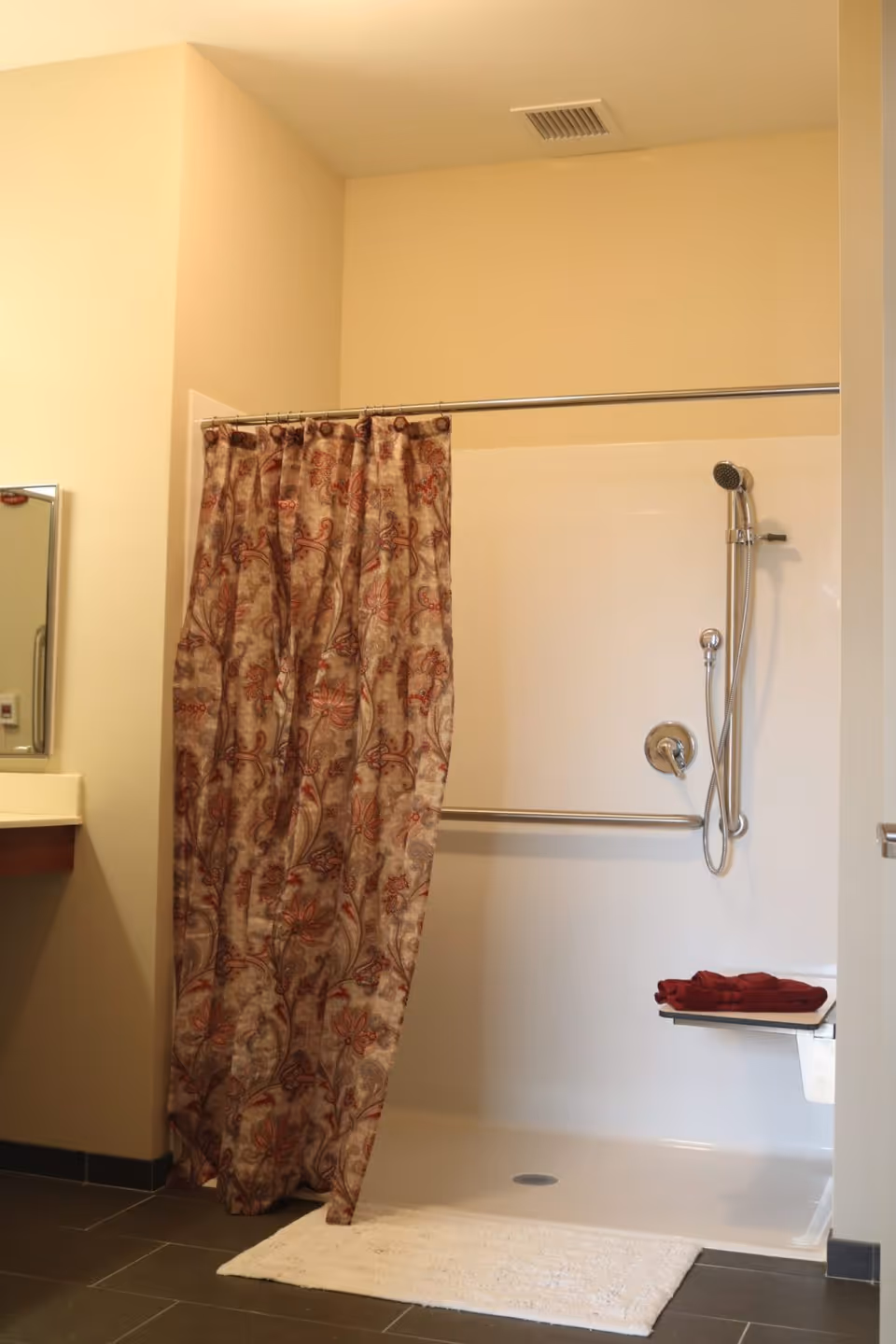 A bathroom shower area with a floral patterned shower curtain partially drawn. The shower has a handheld showerhead attached to a vertical bar and a horizontal grab bar. There is a small fold-down seat with a folded red towel on it. A white bath mat is placed on the dark tiled floor outside the shower.