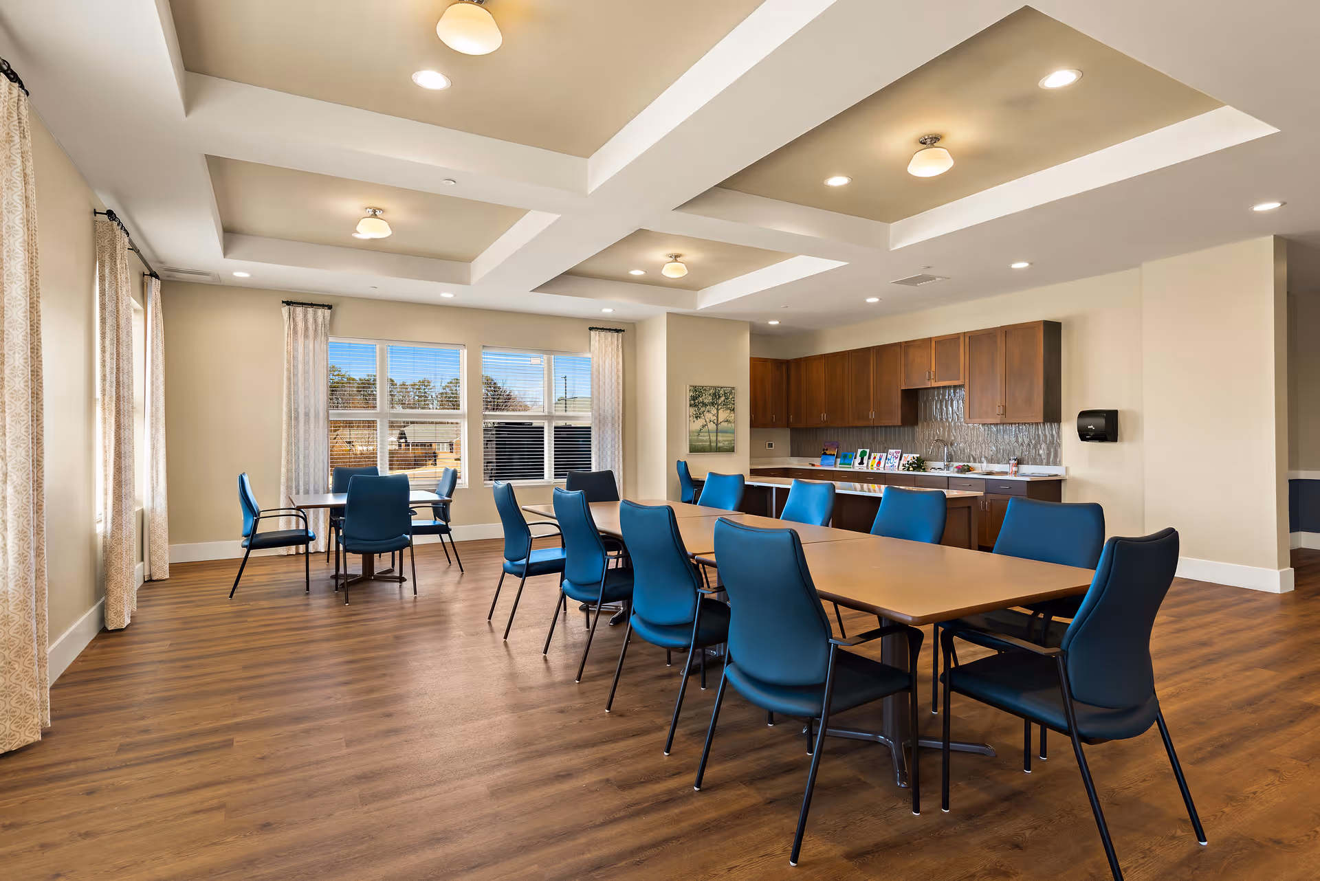 A spacious dining room with a long rectangular table surrounded by blue chairs. The room has large windows with curtains allowing natural light to fill the space. There is a kitchenette area with wooden cabinets and a countertop along one wall. The floor is wood, and the ceiling has recessed lighting and decorative beams.
