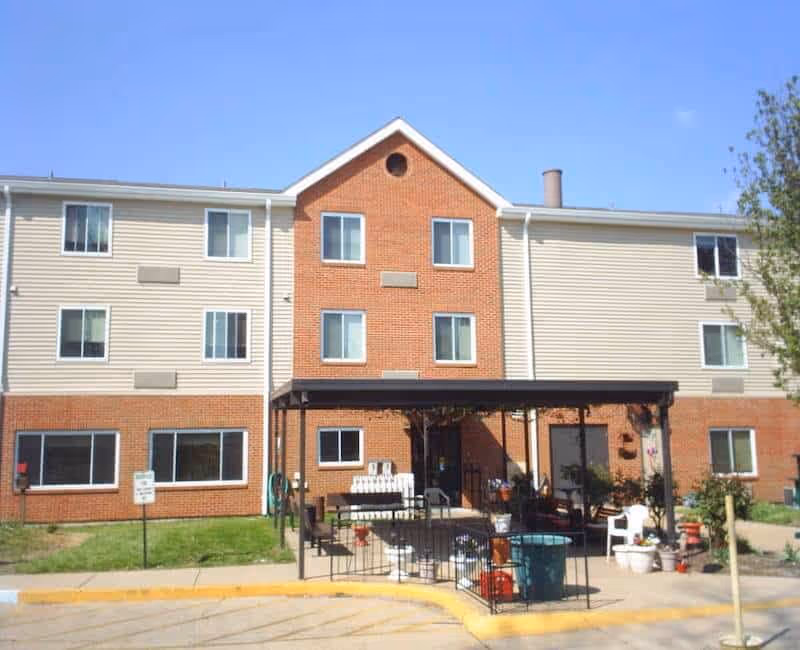 Exterior view of a three-story senior living facility building with a combination of brick and beige siding. The building has multiple windows and a covered entrance area with outdoor seating and potted plants. The sky is clear and blue.