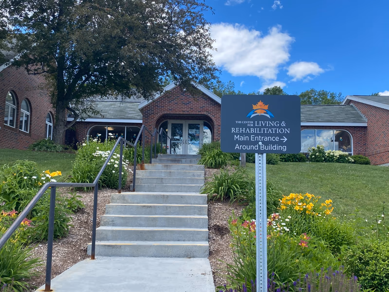 Exterior view of a brick building with a staircase leading up to the entrance. There is a signpost in front of the stairs that reads 'The Center for Living & Rehabilitation Main Entrance Around Building'. The area is landscaped with green grass, flowering plants, and a large tree. The sky is blue with some clouds.