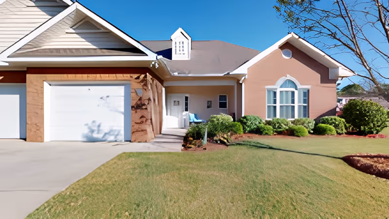 Front exterior of a single-story cottage with an attached garage, manicured lawn, shrubs, and a small covered porch with chairs under a clear blue sky.