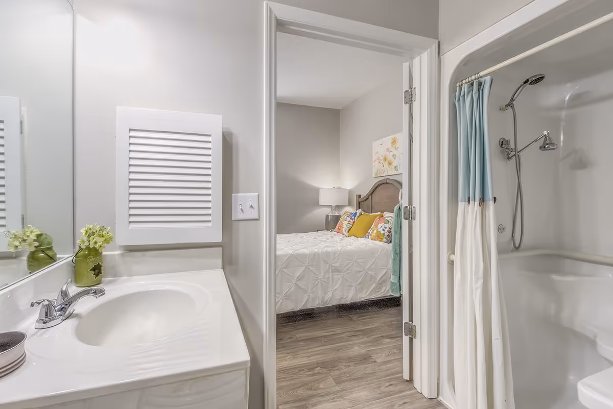 View of a bathroom with a white sink and faucet on the left, a small green vase with flowers on the counter, and a shower with a blue and white curtain on the right. Through an open door, a bedroom is visible with a bed featuring a white comforter, colorful pillows, a wooden headboard, a bedside lamp, and a floral wall decoration.