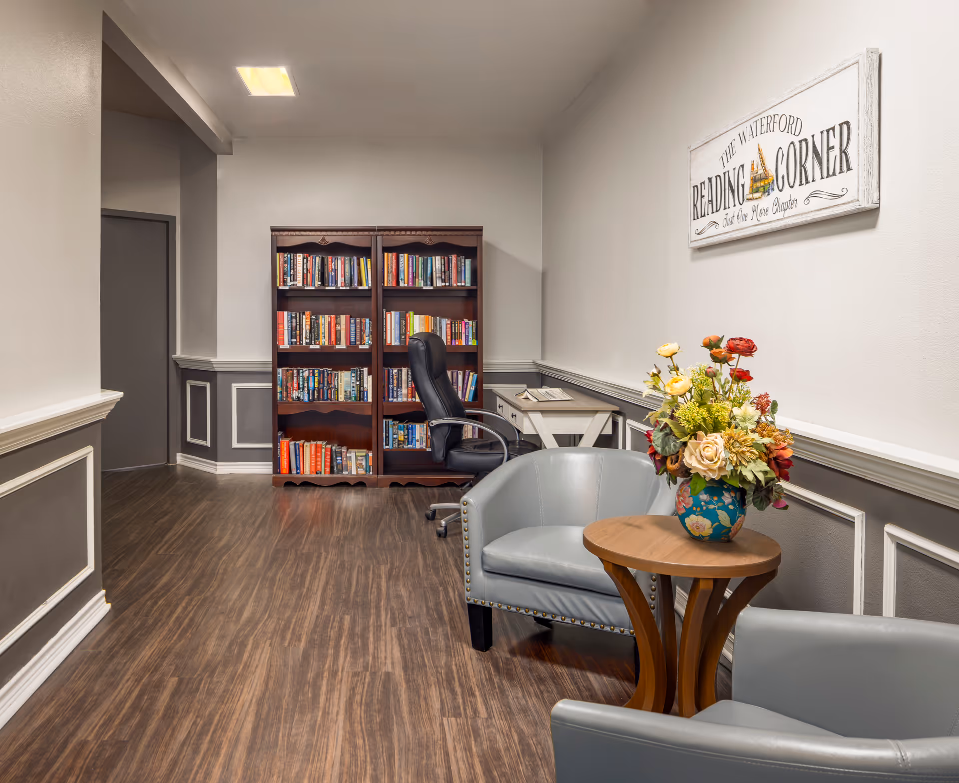 A cozy reading corner with bookshelves, an office chair, two leather armchairs, a small wooden table holding a vase of flowers, and a wall sign that reads "The Waterford Reading Corner."