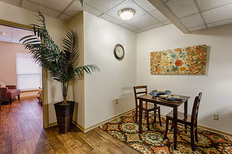 A small dining area with a wooden table set for two, two wooden chairs, a colorful patterned rug underneath, a floral painting on the wall, and a round wall clock. To the left, there is a tall potted plant and an open doorway leading to a room with a window covered by blinds and a red armchair.