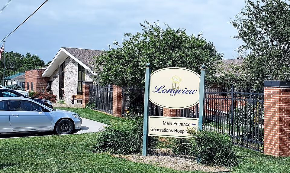 Exterior view of a building with a sign in front that reads 'Longview' with directions to the main entrance and Generations Hospice. There are cars parked on the left side and a black metal fence with brick pillars surrounding the property.