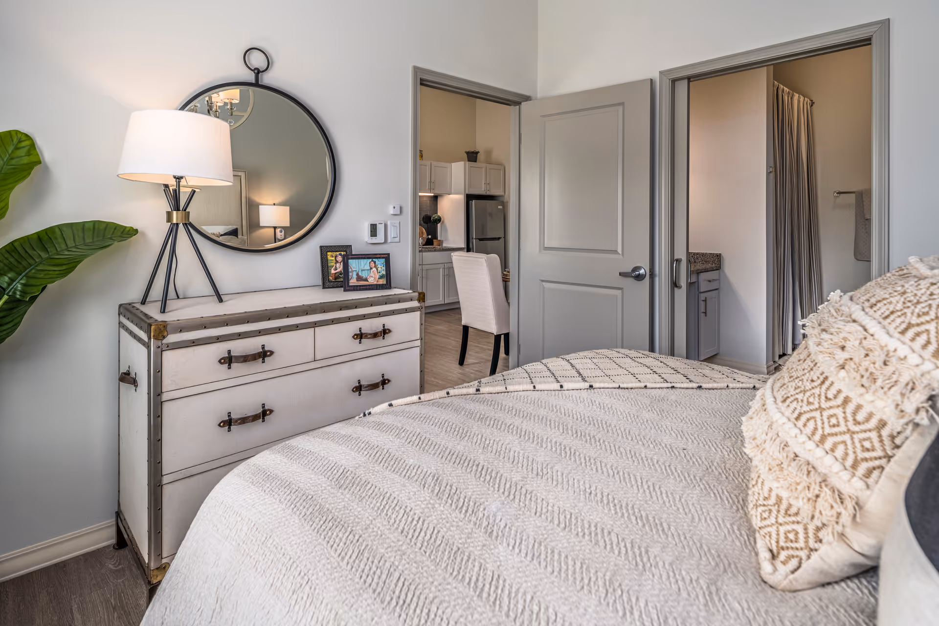 A cozy bedroom with a neatly made bed featuring textured pillows and a patterned blanket. A vintage-style dresser with leather handles holds a modern lamp and two framed photos beneath a round wall mirror. Two open doors reveal a kitchen area with white cabinets and a stainless steel refrigerator, and a bathroom with a granite countertop and a shower curtain.
