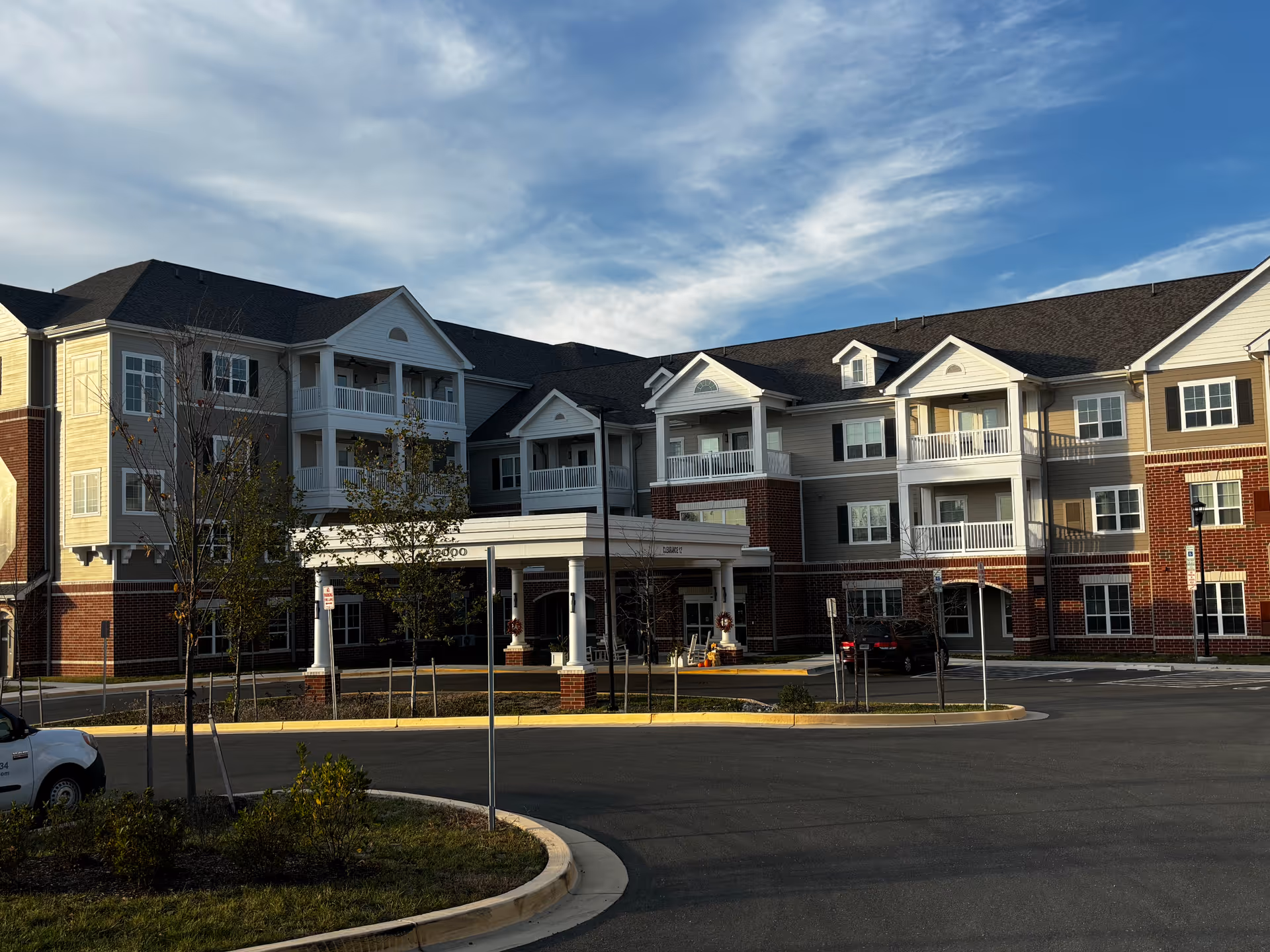 Exterior view of a multi-story senior living facility building with balconies, a covered entrance, and a parking area in front. The building features a combination of brick and siding with white trim, under a partly cloudy sky.