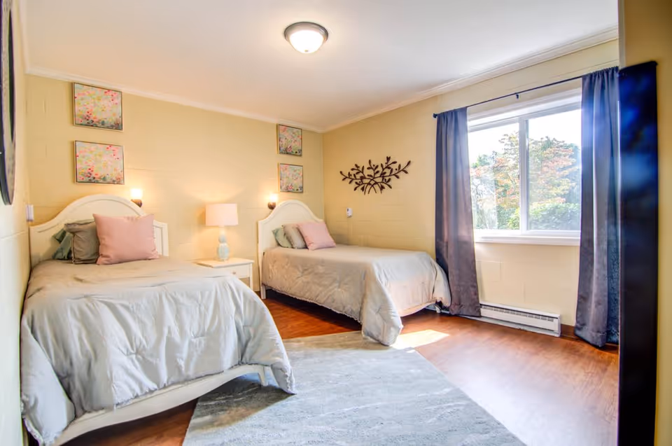 A bright bedroom with two twin beds, each with light gray bedding and pastel pillows. A white nightstand with a lamp is positioned between the beds. The walls are painted a soft yellow and decorated with floral artwork and a metal wall sculpture. A large window with dark curtains lets in natural light, illuminating the wooden floor and a light gray area rug.