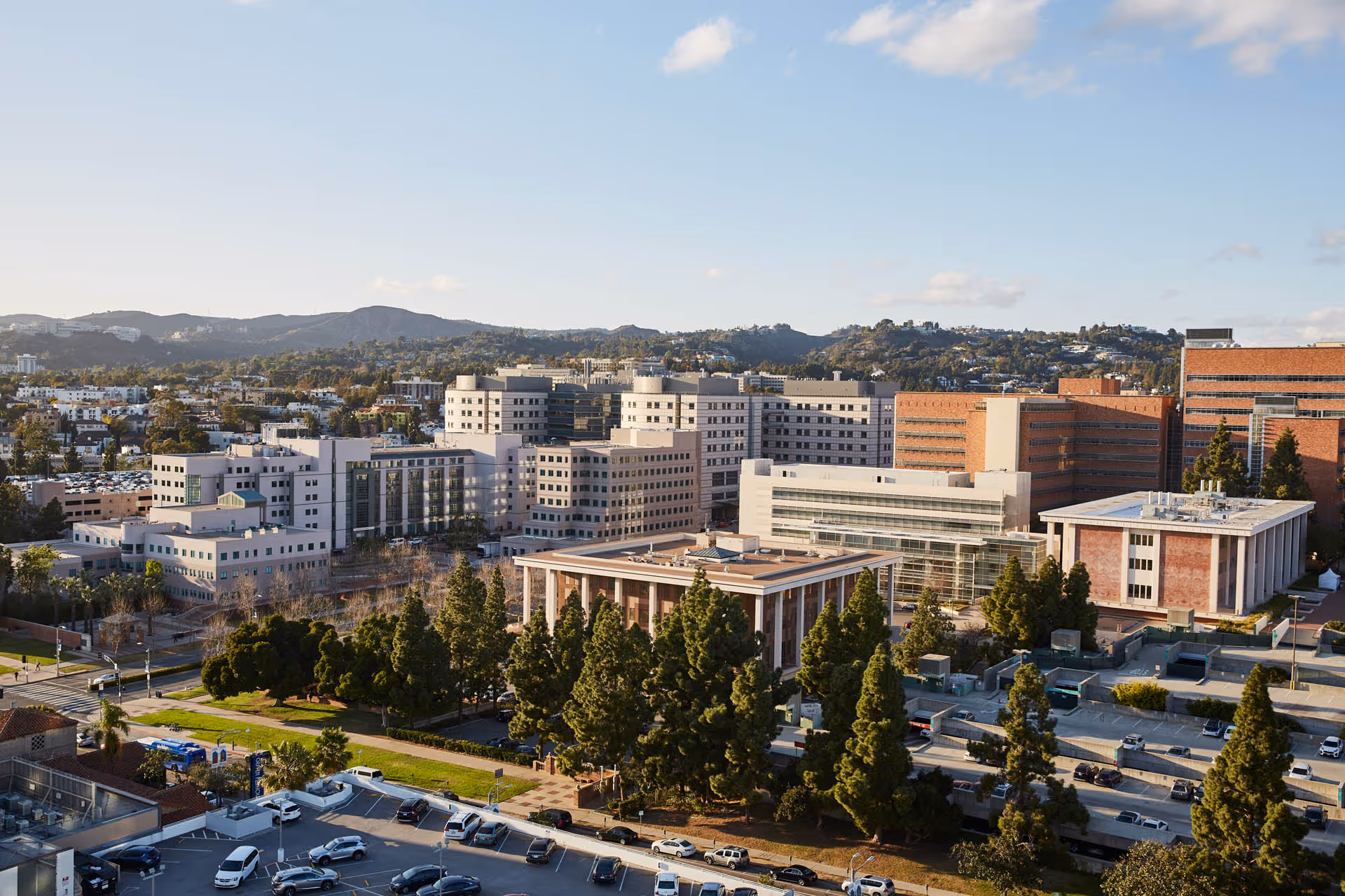A wide exterior view of a cluster of multi-story institutional buildings, surrounding trees, and a parking lot with hills in the background.