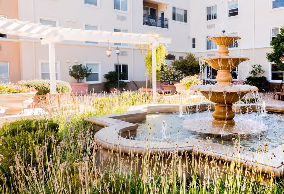 Outdoor courtyard area at Heritage Estates featuring a multi-tiered stone water fountain surrounded by plants and greenery, with a white pergola and beige building with windows in the background.