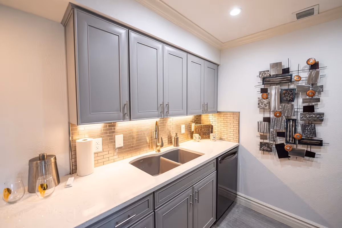 A modern kitchen area with gray cabinets, a white countertop, and a double sink. The backsplash features small rectangular tiles in a metallic finish. Under-cabinet lighting illuminates the countertop. On the counter, there is a paper towel roll, a metal container, two mugs, and a decorative sign that reads 'Good coffee is a pleasure, good friends are a treasure.' A metal abstract wall art piece is mounted on the adjacent white wall.