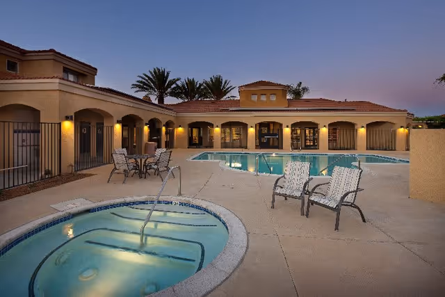 Outdoor pool area at sunset with a hot tub in the foreground, several patio chairs, and a table with chairs. The building surrounding the pool has arched doorways and windows with warm lighting.