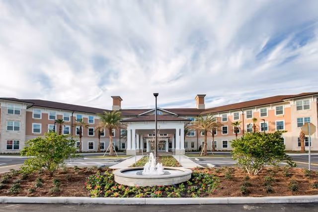 Front exterior view of Canterfield of Ocala, a three-story senior living facility with a central entrance featuring a covered portico, surrounded by landscaped gardens with palm trees and a circular fountain in the foreground under a partly cloudy sky.