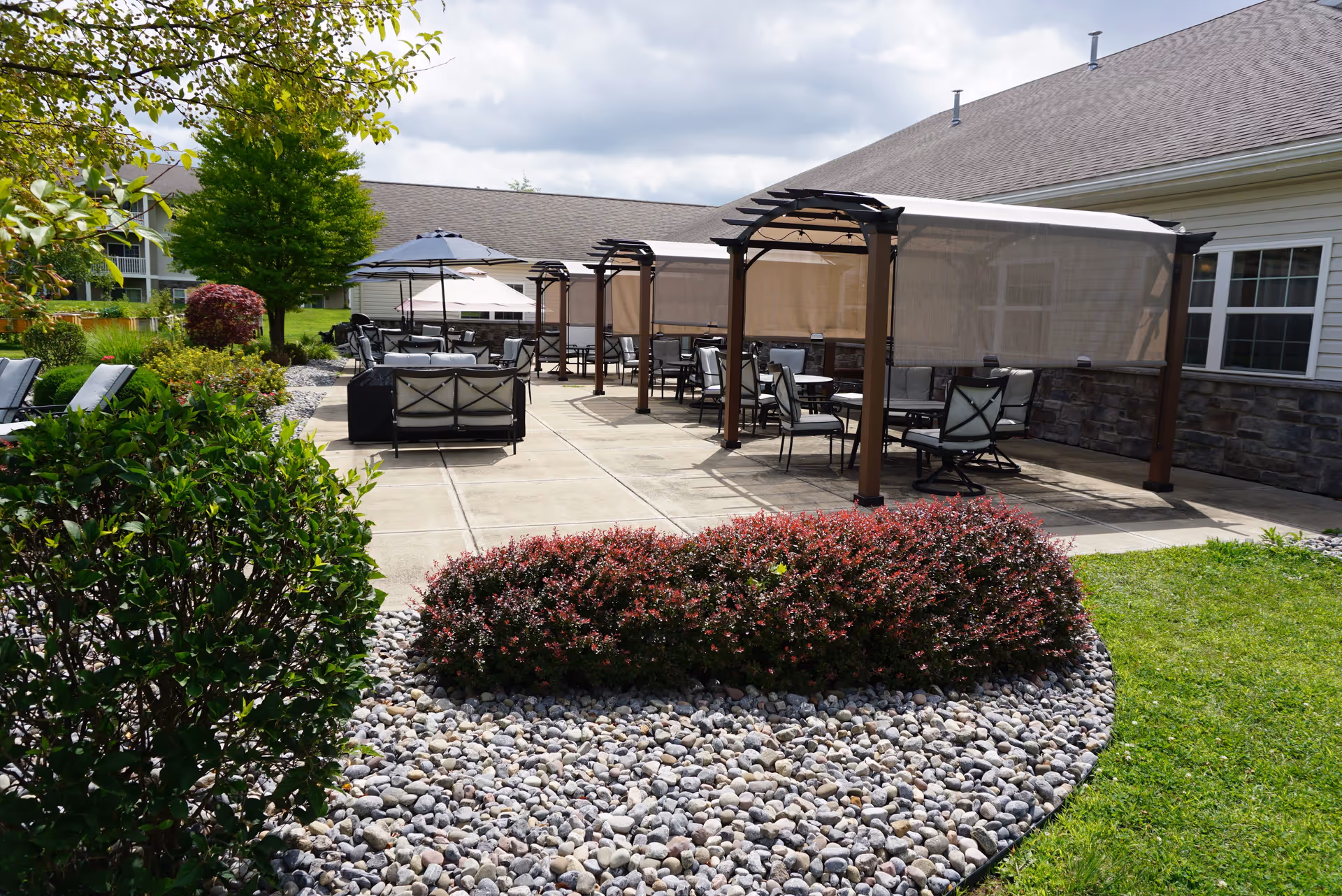 Outdoor patio area at Schuyler Commons with multiple seating arrangements including chairs and tables under pergolas with mesh coverings. The patio is surrounded by landscaped greenery, bushes, and a stone bed, with a building visible on the right side under a partly cloudy sky.