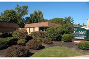 Exterior view of Ambler Extended Care Center building surrounded by well-maintained bushes and trees under a clear blue sky.