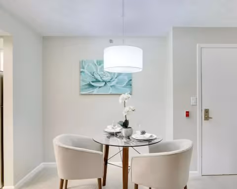 Small dining nook with a round glass table, two upholstered chairs, a hanging pendant light, and wall art.