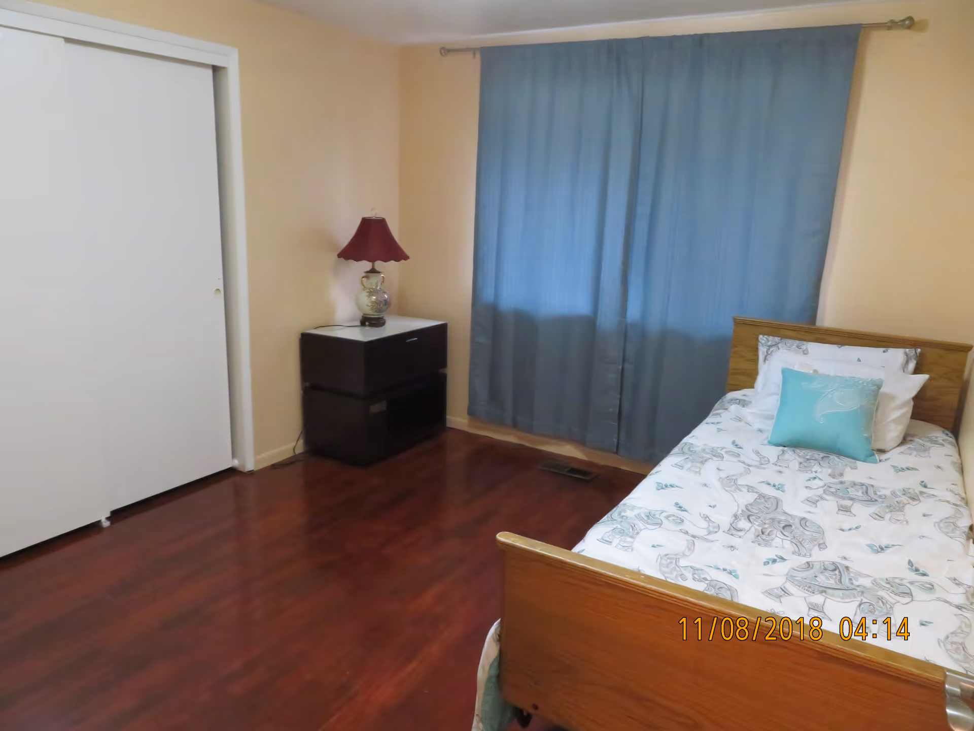 A small bedroom with a single wooden bed covered with white and blue elephant-patterned bedding and a blue pillow. Next to the bed is a dark nightstand with a decorative lamp with a red lampshade. The room has wooden flooring, beige walls, a window covered with blue curtains, and a white sliding closet door.