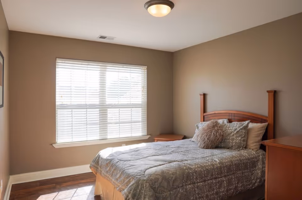 A bedroom with a wooden bed frame, a bed made with patterned bedding and multiple pillows, including a fluffy round pillow. The room has beige walls, a large window with white blinds letting in natural light, a wooden nightstand, and a wooden dresser.