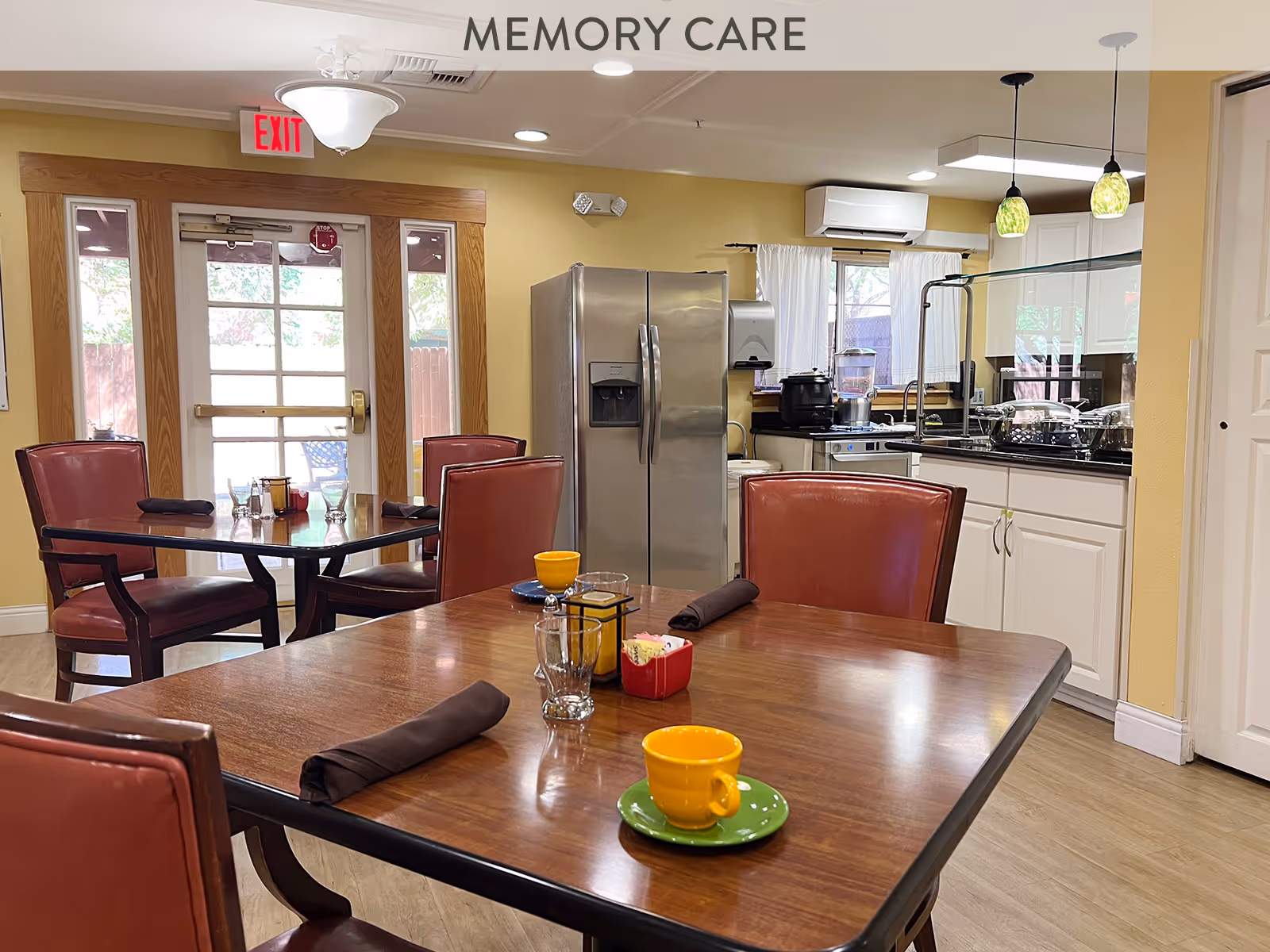 Interior view of a memory care dining area with wooden tables and red cushioned chairs. The tables are set with yellow cups on green saucers, glasses, napkins, and condiments. In the background, there is a kitchen area with white cabinets, a stainless steel refrigerator, and hanging pendant lights. A glass door with an exit sign above it leads outside.