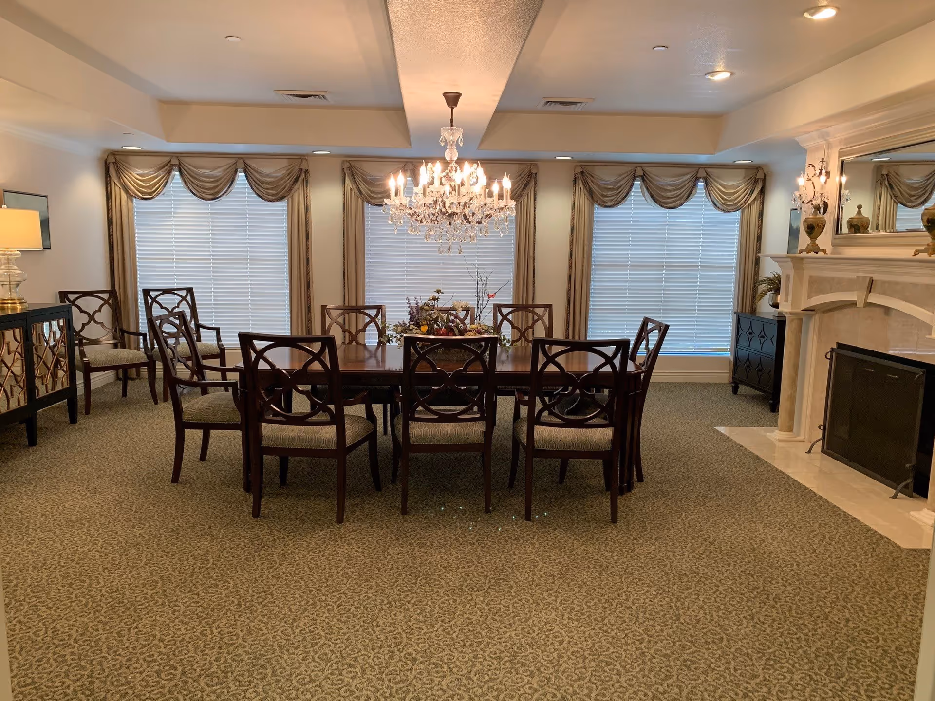 A formal dining room with a large wooden dining table surrounded by eight matching chairs. The room features three large windows with beige curtains and valances, a crystal chandelier hanging above the table, a fireplace with a white mantel on the right, and decorative furniture pieces including a sideboard and a cabinet. The carpet has a subtle patterned design.