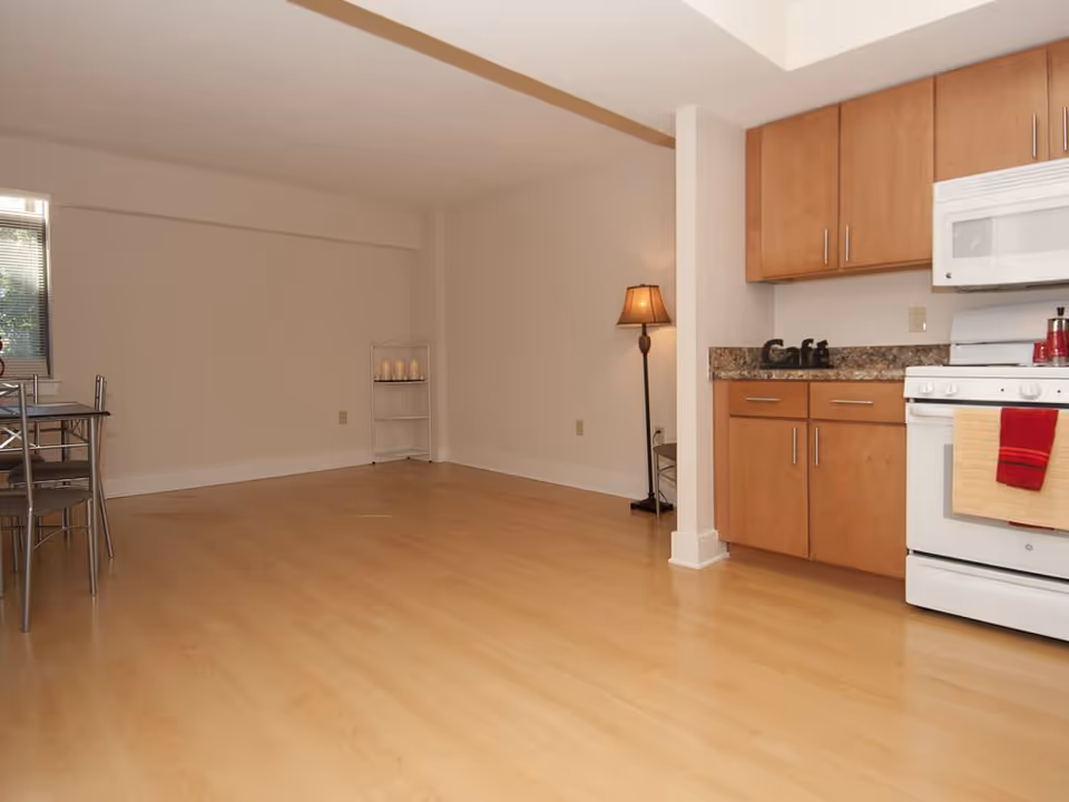 Interior view of a small apartment kitchen and dining area with light wood flooring, wooden cabinets, a white stove with a red and beige towel hanging on the handle, a countertop with a decorative 'Cafe' sign, a floor lamp in the corner, and a metal dining table with chairs near a window with blinds.