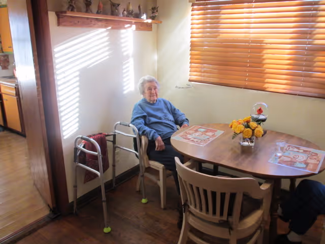 An older adult seated at a small sunlit dining table with a walker beside them and wooden blinds on the window.