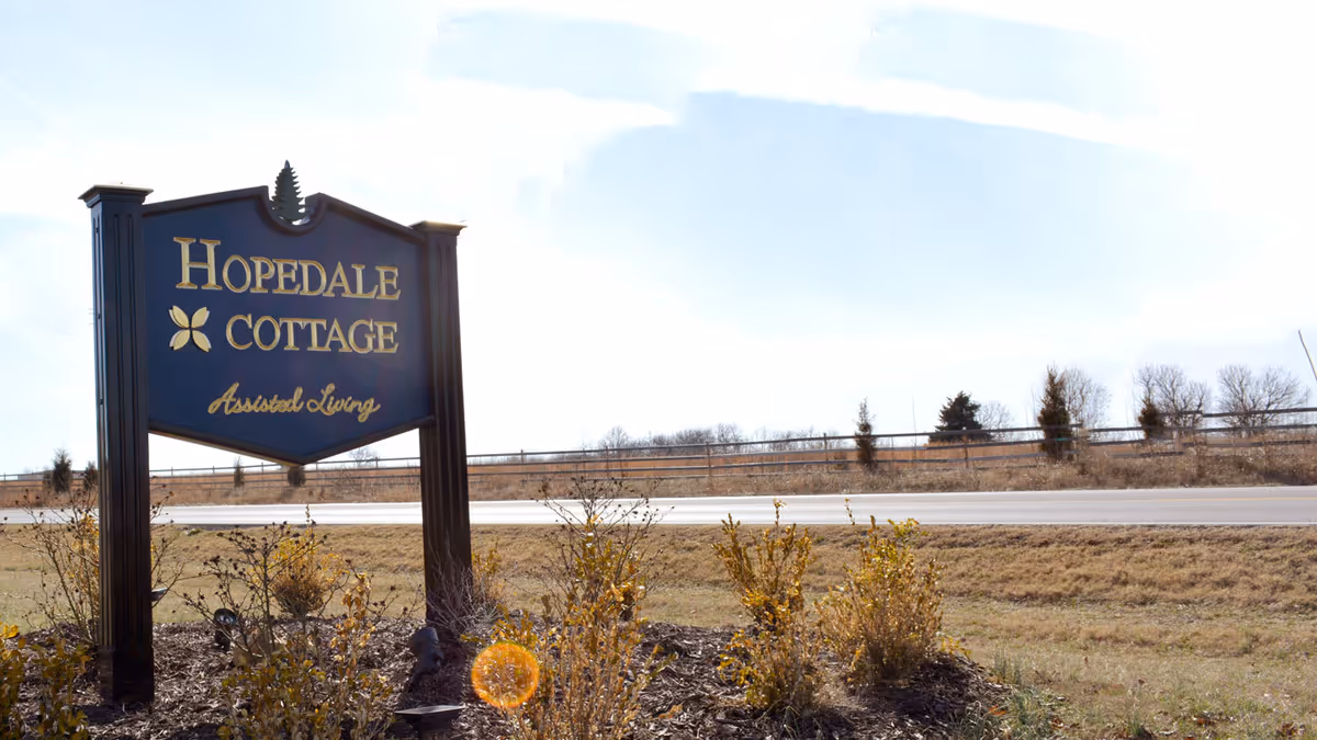 A large dark blue and gold sign for Hopedale Cottage Assisted Living stands in a landscaped area with small bushes and plants, next to a road with a fence and trees in the background under a clear sky.