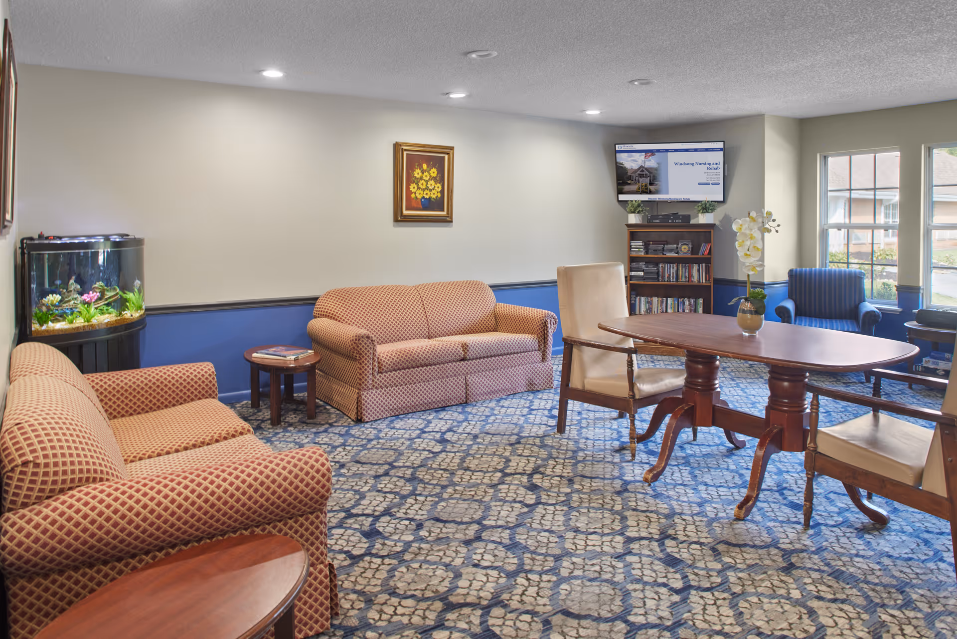 A cozy living room area with two patterned sofas, two beige armchairs around a wooden table, a blue armchair near windows, a small bookshelf with a TV mounted above it, a fish tank in the corner, and a framed painting of sunflowers on the wall.