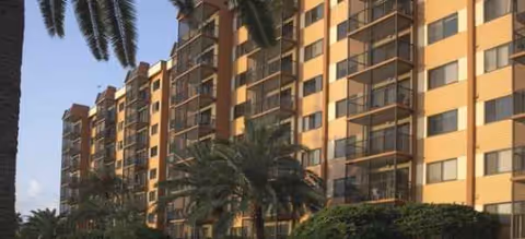 Exterior view of a tall tan senior living building with balconies and palm trees.