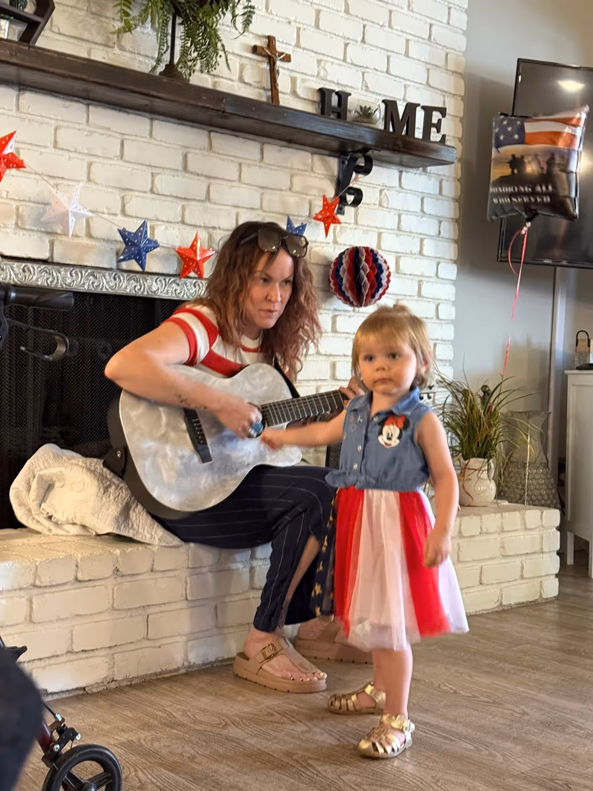 A woman sitting on a white brick hearth playing a guitar while a young girl in a red, white, and blue dress stands nearby. The background features a white brick wall with patriotic star decorations, a wooden shelf with the word HOME and a cross, and a balloon with an American flag design.