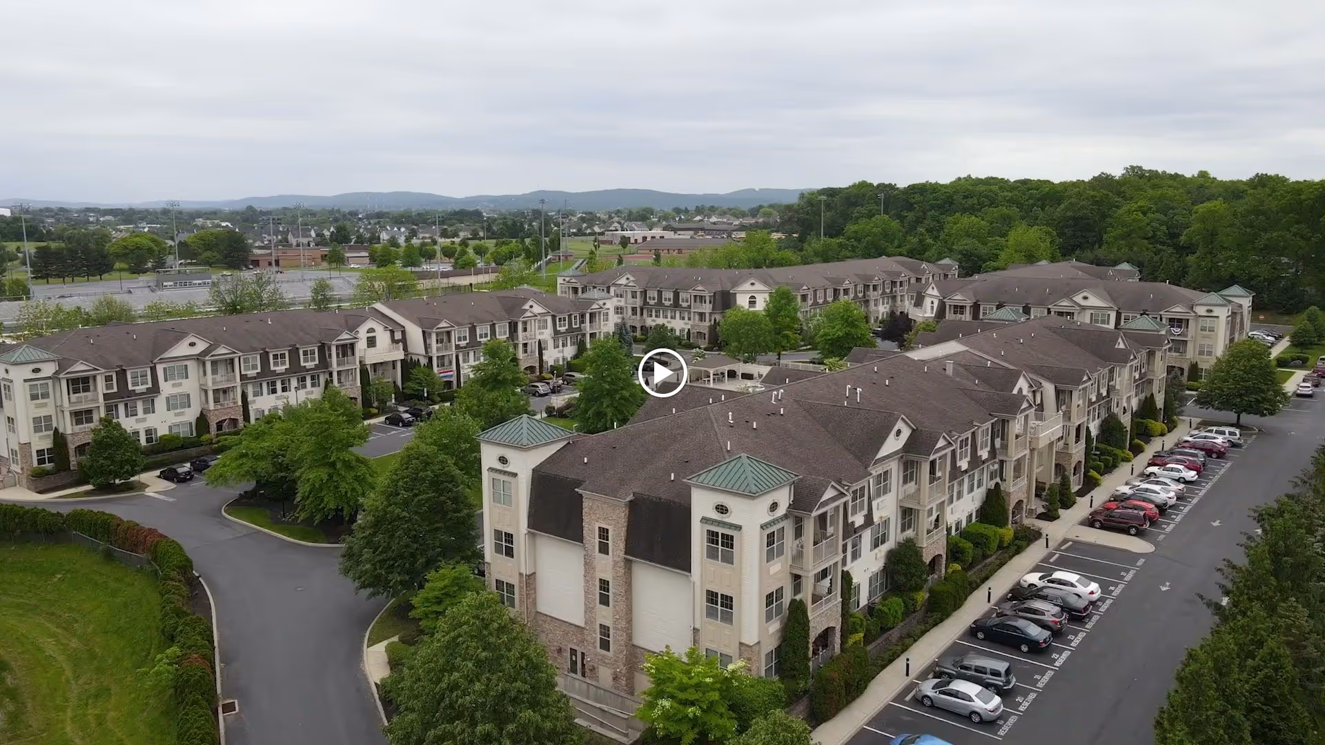Aerial view of a multi-building senior living complex with three-story beige residential buildings, trees and a parking lot with cars.