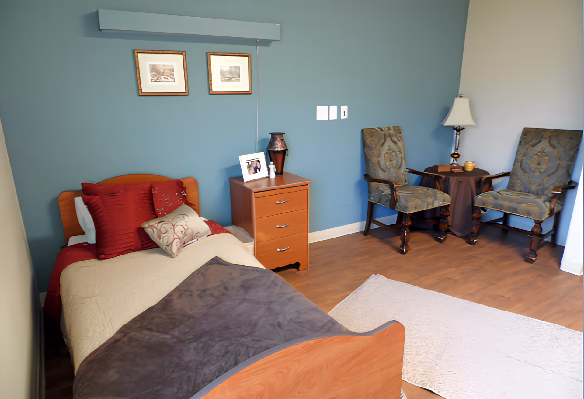 A cozy bedroom in a senior living facility with a single wooden bed adorned with red and beige pillows and blankets. Next to the bed is a wooden nightstand with a framed photo, a decorative vase, and a small bottle. Two upholstered armchairs with a small round table and a lamp are positioned against a blue wall. The floor is wooden with a light-colored rug near the bed.