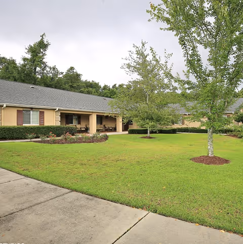 Single-story senior living building with a covered porch facing a green lawn and young trees under an overcast sky.