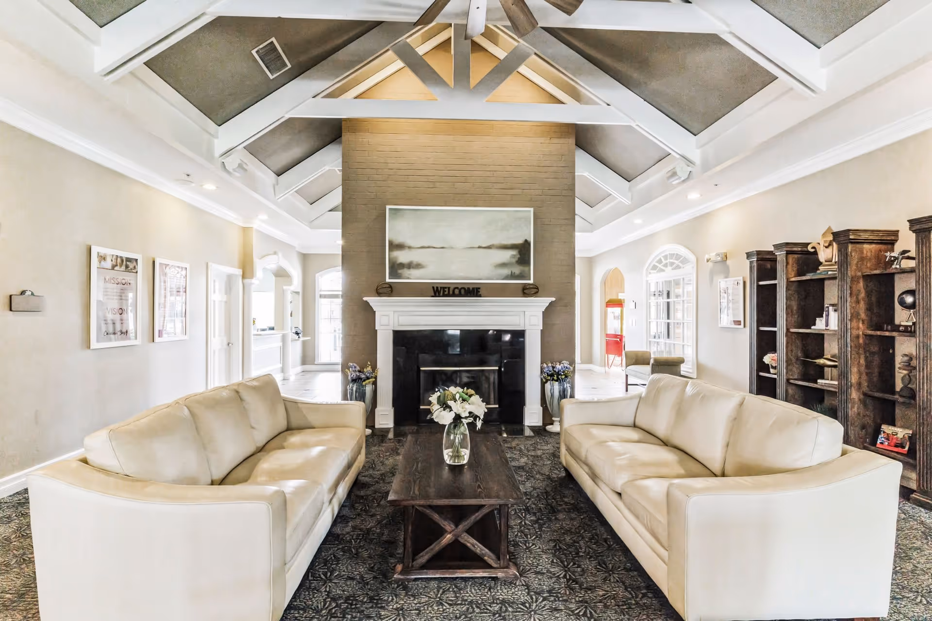 A bright and spacious living room area with two cream-colored sofas facing each other across a dark wooden coffee table with a vase of white flowers. The room features a high vaulted ceiling with exposed white beams, a central fireplace with a 'WELCOME' sign on the mantel, and a large painting above it. There are decorative vases with flowers on either side of the fireplace, framed pictures on the walls, and wooden bookshelves on the right side of the room.