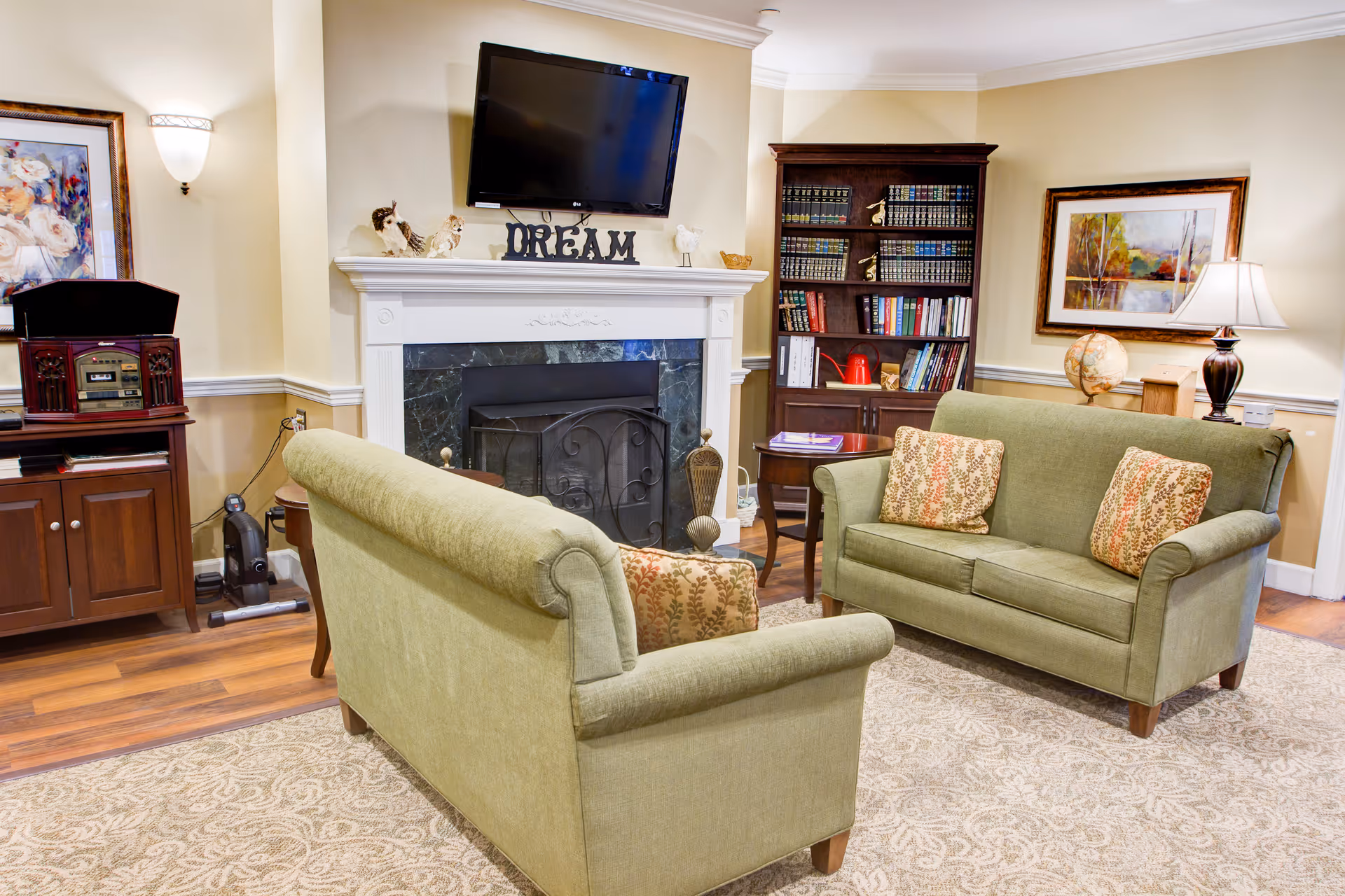 Cozy common living room with two green sofas facing a fireplace and TV, a bookshelf, and side tables with lamps.