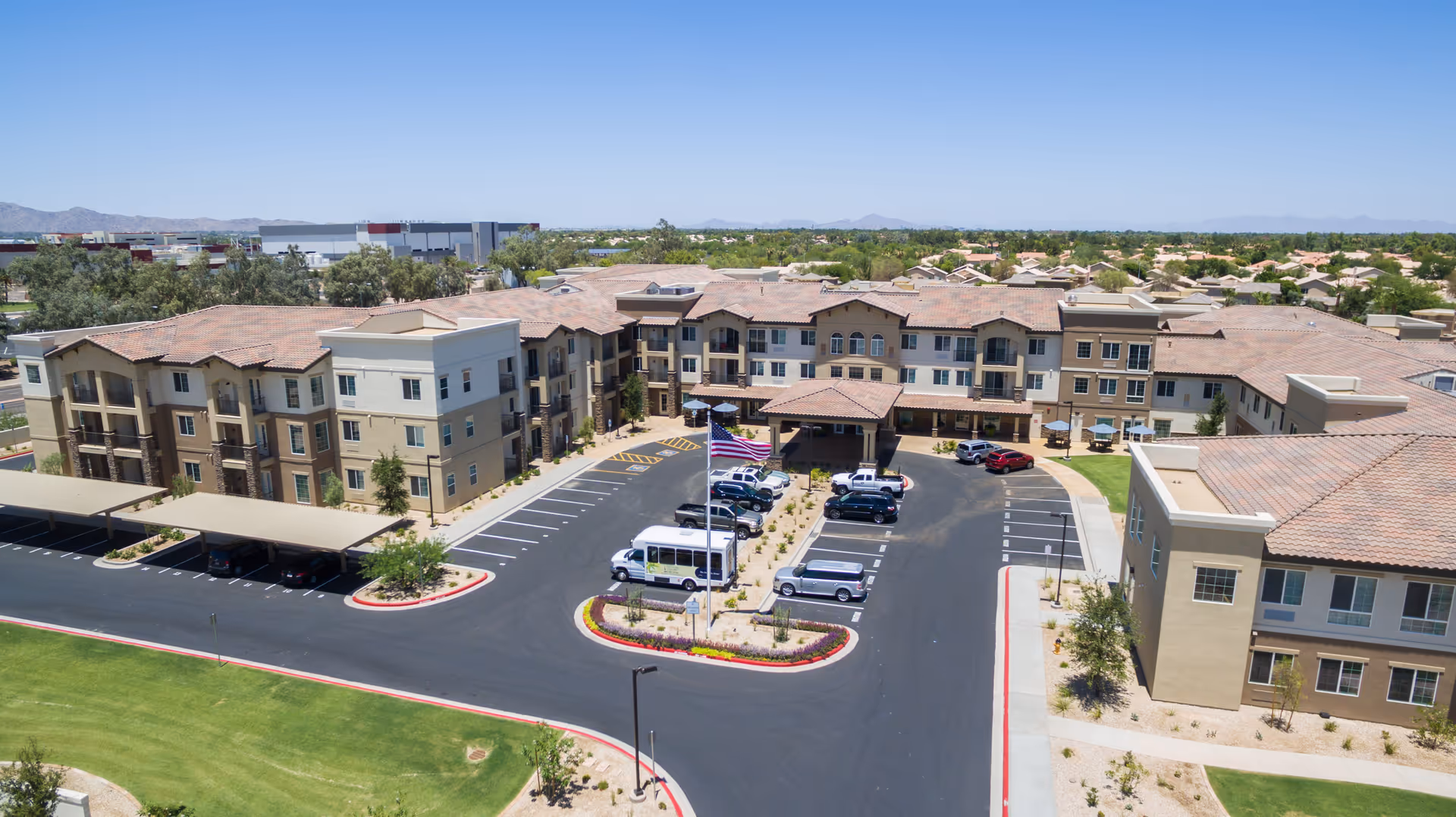 Aerial view of The Enclave at Chandler Senior Living facility showing a large U-shaped building with multiple floors, a central parking lot with several cars and a shuttle van, covered parking areas, landscaped greenery, and a clear blue sky in the background.