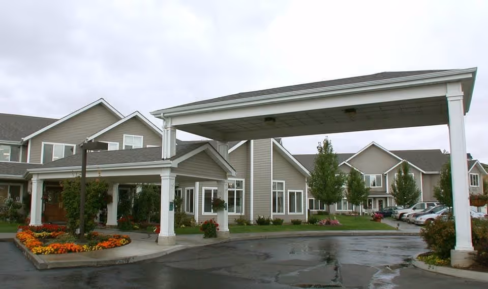 Exterior view of Bishop Place Senior Living facility showing a covered entrance with white pillars, a driveway, landscaped flower beds, and multiple connected beige buildings with white trim under an overcast sky.
