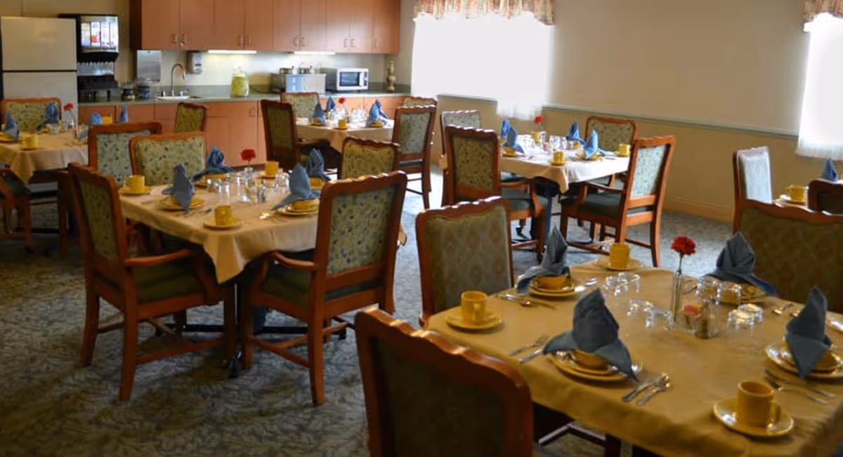 A dining room in a senior living facility with several tables set with yellow cups, plates, silverware, and blue folded napkins. The room has upholstered wooden chairs, carpeted floor, and a kitchenette area with cabinets, a refrigerator, microwave, and coffee machine in the background. Natural light comes through windows with sheer curtains.