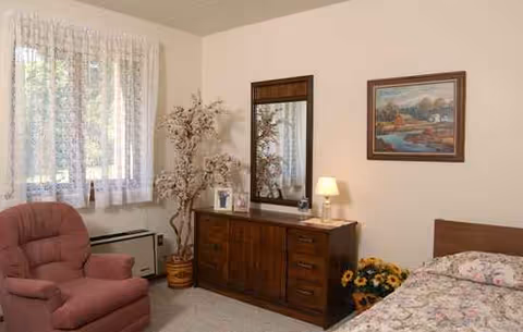 Cozy retirement home bedroom with bed, dresser, lamp, pink recliner, and lace-curtained window.