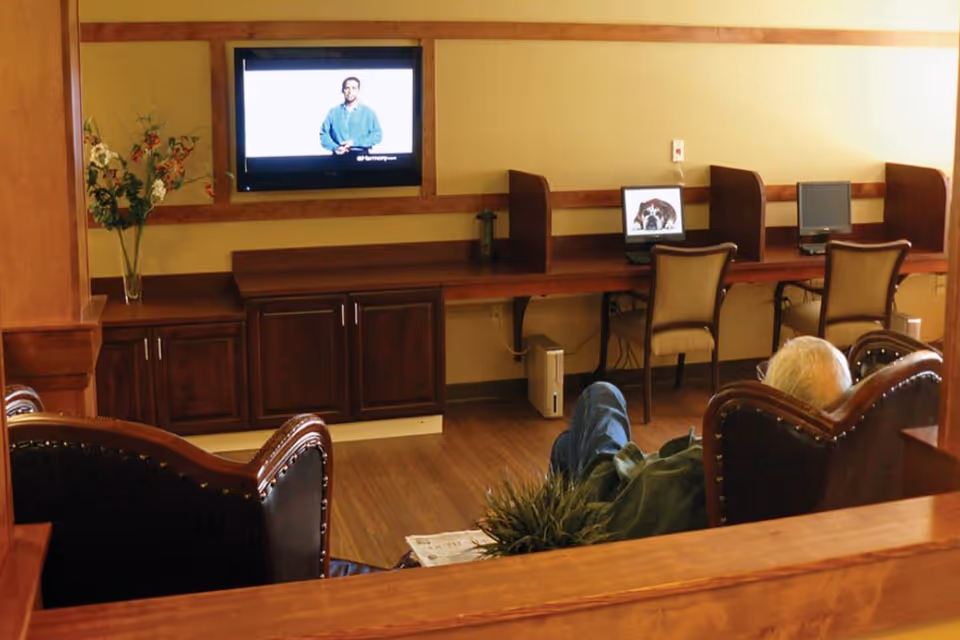 A cozy common area with wooden furniture and flooring. There are two upholstered chairs facing a wall-mounted TV showing a man in a blue shirt. Along the wall, there is a long wooden desk with three computer stations, one displaying a dog image. A vase with flowers is placed on a cabinet under the TV.