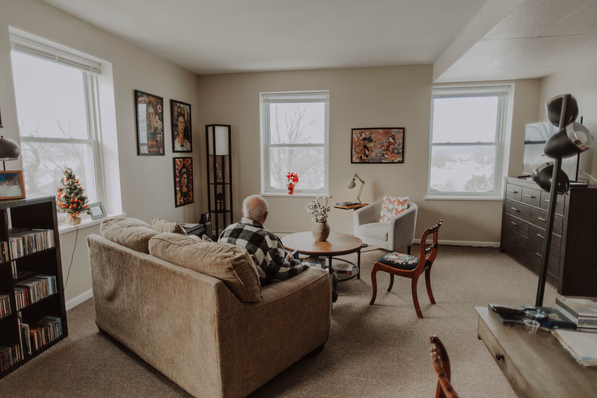 A cozy living room with a person seated on a sofa facing three large windows, a coffee table, chairs, and wall art.