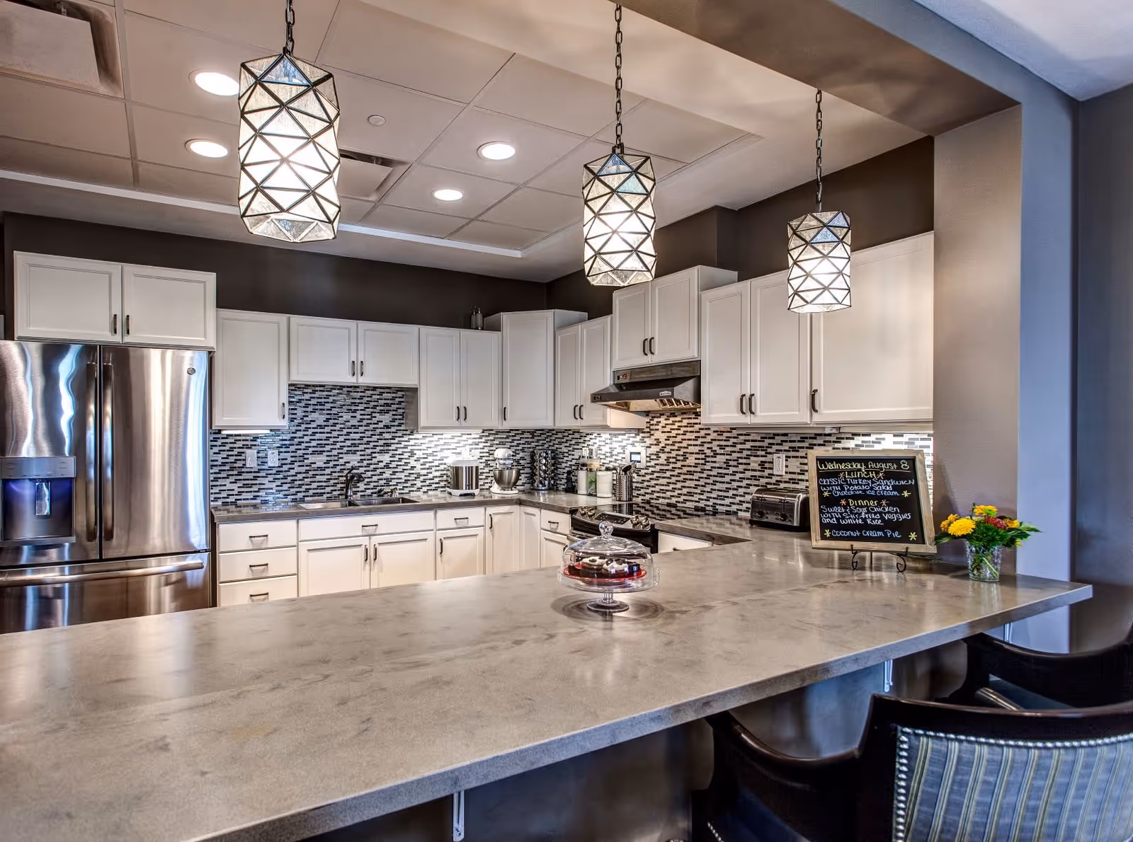 Modern kitchen with white cabinets, a stainless steel refrigerator, a tiled backsplash, and three geometric pendant lights hanging over a large marble countertop island with chairs. A small chalkboard menu and a vase with flowers are placed on the countertop.